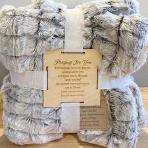 May include: A soft, gray and white faux fur blanket with a wooden tag that reads "Praying For You." The blanket is tied with a white ribbon. The tag includes a message of comfort and prayer.
