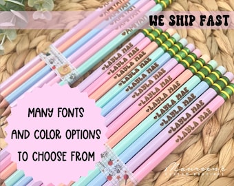 May include: A bundle of pastel colored pencils with personalized names in a gold font. The pencils have green and yellow stripes. The text "MANY FONTS AND COLOR OPTIONS TO CHOOSE FROM" is visible in the image. The text "WE SHIP FAST" is also visible in the image.