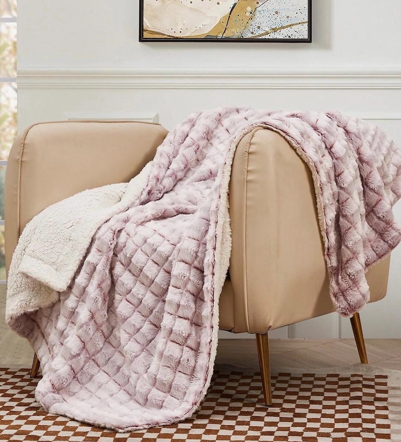 May include: A plush, lavender throw blanket with a diamond pattern is draped over a beige armchair. The blanket has a soft, textured surface and a cream-colored, fuzzy underside. The chair has gold-colored legs and sits on a checkered rug.