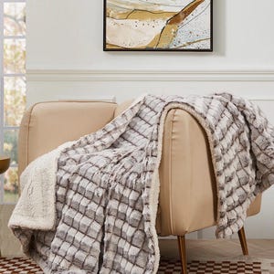 May include: A plush, checkered gray and white blanket draped over a beige armchair. The blanket has a soft, textured appearance with a white, fluffy underside. The room features a painting and a checkered rug.