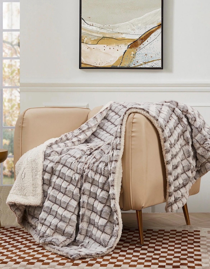 May include: A plush, checkered gray and white blanket draped over a beige armchair. The blanket has a soft, textured appearance with a white, fluffy underside. The room features a painting and a checkered rug.