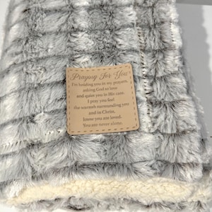 May include: A gray and white faux fur throw blanket with a brown tag attached. The tag has the text "Praying For You" and a message about holding someone in prayers and asking God to love and quiet them in His care. The tag also says "I pray you feel the warmth surrounding you and in Christ, know you are loved. You are never alone."
