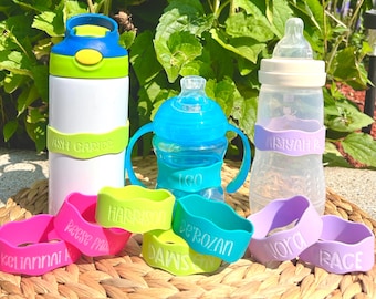 May include: Eight colorful silicone bottle labels with names printed on them. The labels are on a woven straw mat.  The labels are for water bottles, sippy cups, and baby bottles.