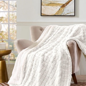 May include: A plush, white throw blanket draped over a beige armchair. The blanket has a textured, quilted pattern and a soft, fluffy appearance. The room features a window, artwork, and a small side table.