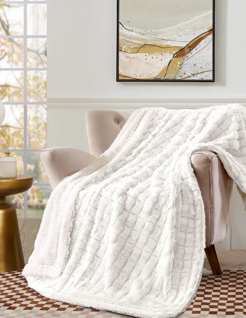 May include: A plush, white throw blanket draped over a beige armchair. The blanket has a textured, quilted pattern and a soft, fluffy appearance. The room features a window, artwork, and a small side table.