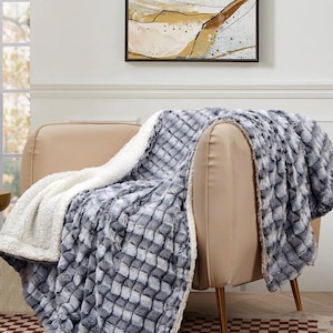 May include: A plush, reversible blanket draped over a beige armchair. One side features a textured, gray and white diamond pattern, while the other is a soft, white sherpa material. The blanket adds a cozy touch to the room.
