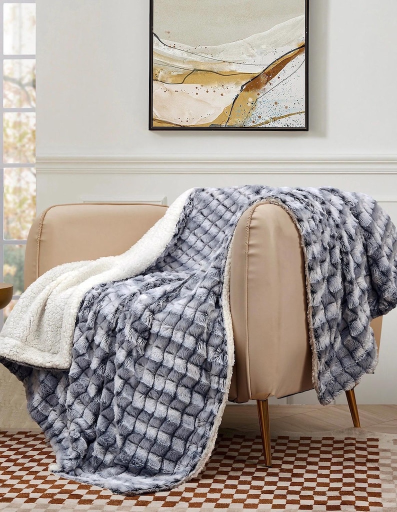 May include: A plush, reversible blanket draped over a beige armchair. One side features a textured, gray and white diamond pattern, while the other is a soft, white sherpa material. The blanket adds a cozy touch to the room.