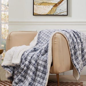 May include: A soft, plush blanket draped over a beige armchair. The blanket features a textured, quilted pattern in shades of blue and gray, with a white, fluffy sherpa lining. The blanket is designed for comfort and warmth.