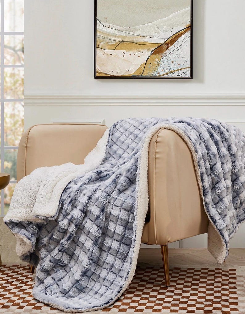 May include: A soft, plush blanket draped over a beige armchair. The blanket features a textured, quilted pattern in shades of blue and gray, with a white, fluffy sherpa lining. The blanket is designed for comfort and warmth.