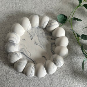 May include: A white and gray marble trinket dish with a scalloped edge. The dish is round and has a smooth, polished surface.
