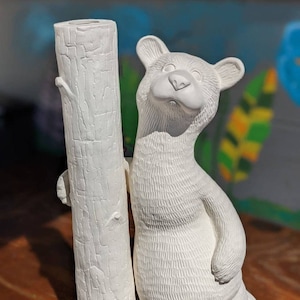 May include: A white ceramic bear figurine leaning against a tree trunk. The bear has a friendly expression and textured fur. The tree trunk is also white and has a detailed bark pattern. This decorative item is suitable for home decor.