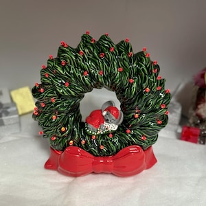 Può includere: Una ghirlanda natalizia in ceramica con finitura verde scuro lucida e piccoli accenti rossi. Un piccolo topo grigio con un cappellino rosso dorme al centro della ghirlanda, decorata con un grande fiocco rosso a tinta unita alla base.