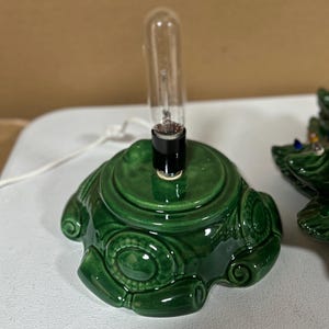 May include: A green ceramic Christmas tree base with a clear light bulb. The base has a decorative, embossed design and a black socket. The base is on a white surface.