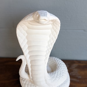 Może przedstawiać: Biała rzeźba kobry o szczegółowej, teksturowanej powierzchni. Wąż przedstawiony jest w uderzającej, wyprostowanej pozie, z rozłożoną kapturem. Rzeźba umieszczona jest na neutralnym tle, podkreślając skomplikowany wzór kobry.