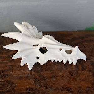May include: A white dragon skull mask with detailed features, including horns, eye sockets, and sharp teeth. The mask is made of a smooth, matte material and is designed to be worn or displayed as a decorative item.