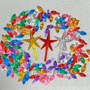 May include: A collection of colorful Christmas light bulbs and star-shaped tree toppers. The light bulbs are in various colors, including red, green, yellow, blue, pink, and purple. The star toppers are gold, red, and clear.