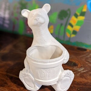 Op de afbeelding: Witte keramische berenpot met een emmer als buik. De beer zit op een houten oppervlak.