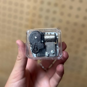 May include: A clear, rectangular music box with visible internal mechanisms. The box features a black winding mechanism and a silver-colored cylinder with small pins. The music box is held in a hand, showcasing its compact size and intricate design.