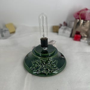 May include: A dark green ceramic lamp base featuring a raised holly leaf and berry design. The circular base has a central black socket holding a clear, tubular incandescent light bulb. A white electrical cord extends from the back of the base.
