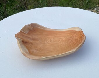 May include: A handcrafted wooden bowl with a natural, organic shape. The bowl is made from light brown wood, showcasing the wood grain. The bowl is on a white surface, with green grass in the background. This decorative bowl is perfect for serving.