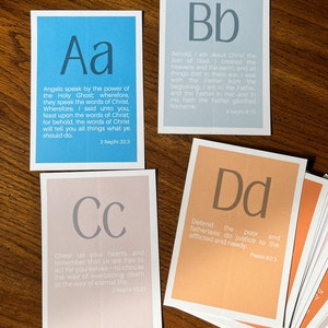 May include: A set of alphabet flashcards with a scripture quote on each card. The cards are in different colours, including blue, light blue, pink, and orange. The letters are in a large, bold font. The quotes are from the Book of Mormon and the Bible.