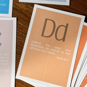May include: A set of alphabet flashcards with a peach background and brown lettering. Each card features a letter of the alphabet and a corresponding Bible verse. The card shown is for the letter "D" and the verse is "Defend the poor and fatherless: do justice to the afflicted and needy." Psalm 82:3.