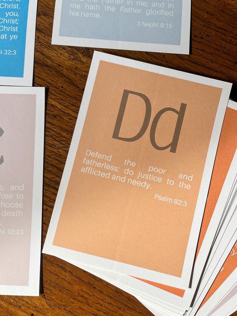 May include: A set of alphabet flashcards with a peach background and brown lettering. Each card features a letter of the alphabet and a corresponding Bible verse. The card shown is for the letter "D" and the verse is "Defend the poor and fatherless: do justice to the afflicted and needy." Psalm 82:3.