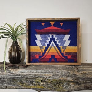 May include: A framed wall tapestry with a geometric design in shades of blue, red, orange, and yellow. The tapestry is framed in a dark wood frame.