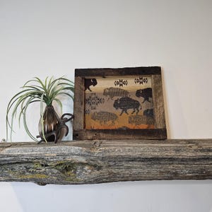 May include: A framed textile art piece featuring a rustic design with bison silhouettes. The artwork is set in a reclaimed wood frame. A decorative plant and metal sculpture are on a wooden shelf.