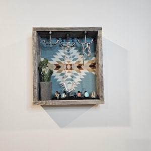 May include: A rustic wooden jewelry display case with a Southwestern-style patterned fabric backing. The case features three metal hooks for hanging necklaces, a small potted cactus, and a black tray holding several rings. The color palette includes shades of gray, blue, and brown.