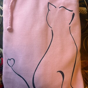 Op de afbeelding: Een lichtroze hoodie met een zwarte omtrek van een zittende kat, met een hartje aan het einde van de staart. De hoodie heeft een trekkoord en een capuchon.