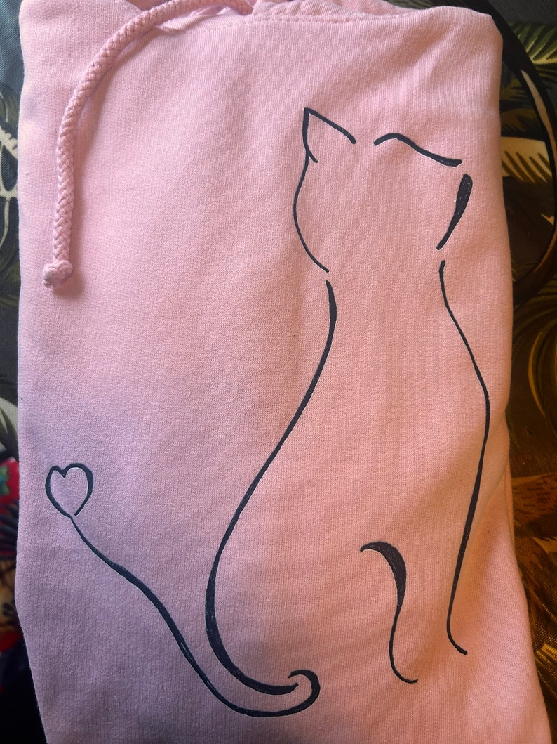 Op de afbeelding: Een lichtroze hoodie met een zwarte omtrek van een zittende kat, met een hartje aan het einde van de staart. De hoodie heeft een trekkoord en een capuchon.