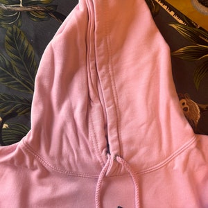Op de afbeelding: Een lichtroze hoodie met een trekkoord en een capuchon. De capuchon is omhoog en het trekkoord is zichtbaar. De hoodie heeft een eenvoudig ontwerp, met een zwarte afbeelding op de voorkant. De stof ziet er zacht en comfortabel uit.