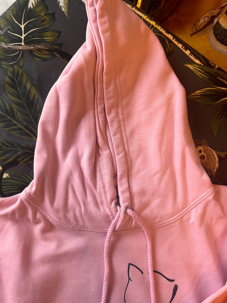Op de afbeelding: Een lichtroze hoodie met een trekkoord en een capuchon. De capuchon is omhoog en het trekkoord is zichtbaar. De hoodie heeft een eenvoudig ontwerp, met een zwarte afbeelding op de voorkant. De stof ziet er zacht en comfortabel uit.