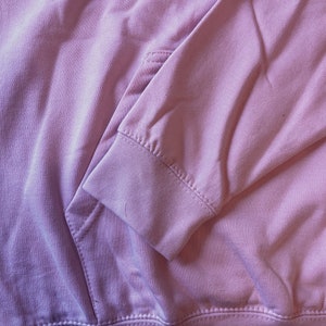 Op de afbeelding: Close-up van een lichtroze hoodie. De afbeelding toont de details van de zak, mouw en zoom. De stof lijkt zacht en de stiksels zijn zichtbaar. De hoodie is effen van kleur en heeft een casual stijl.