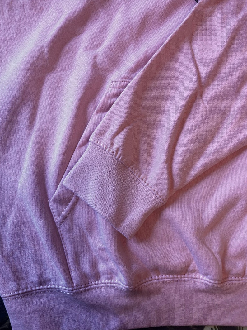 Op de afbeelding: Close-up van een lichtroze hoodie. De afbeelding toont de details van de zak, mouw en zoom. De stof lijkt zacht en de stiksels zijn zichtbaar. De hoodie is effen van kleur en heeft een casual stijl.