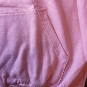 Op de afbeelding: Close-up van een lichtroze hoodie met een kangoeroezak. De zak is gestikt met zichtbare naden en de stof heeft een zachte, licht getextureerde uitstraling. De hoodie is gemaakt van een dik, comfortabel materiaal.