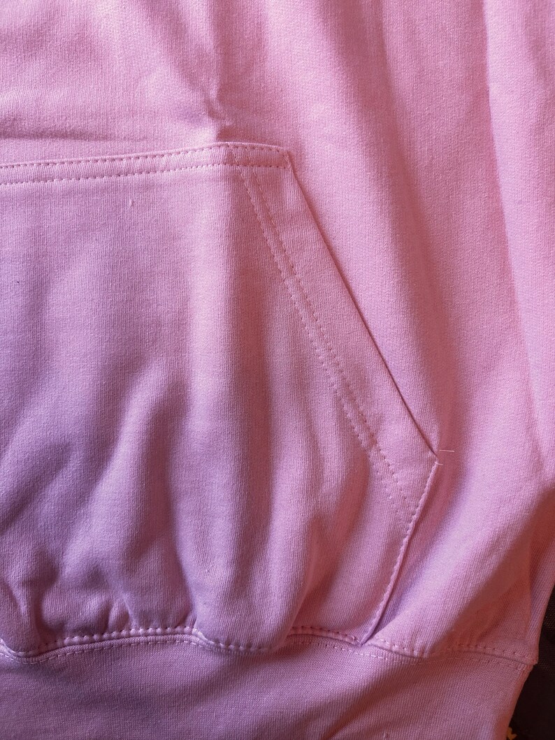 Op de afbeelding: Close-up van een lichtroze hoodie met een kangoeroezak. De zak is gestikt met zichtbare naden en de stof heeft een zachte, licht getextureerde uitstraling. De hoodie is gemaakt van een dik, comfortabel materiaal.