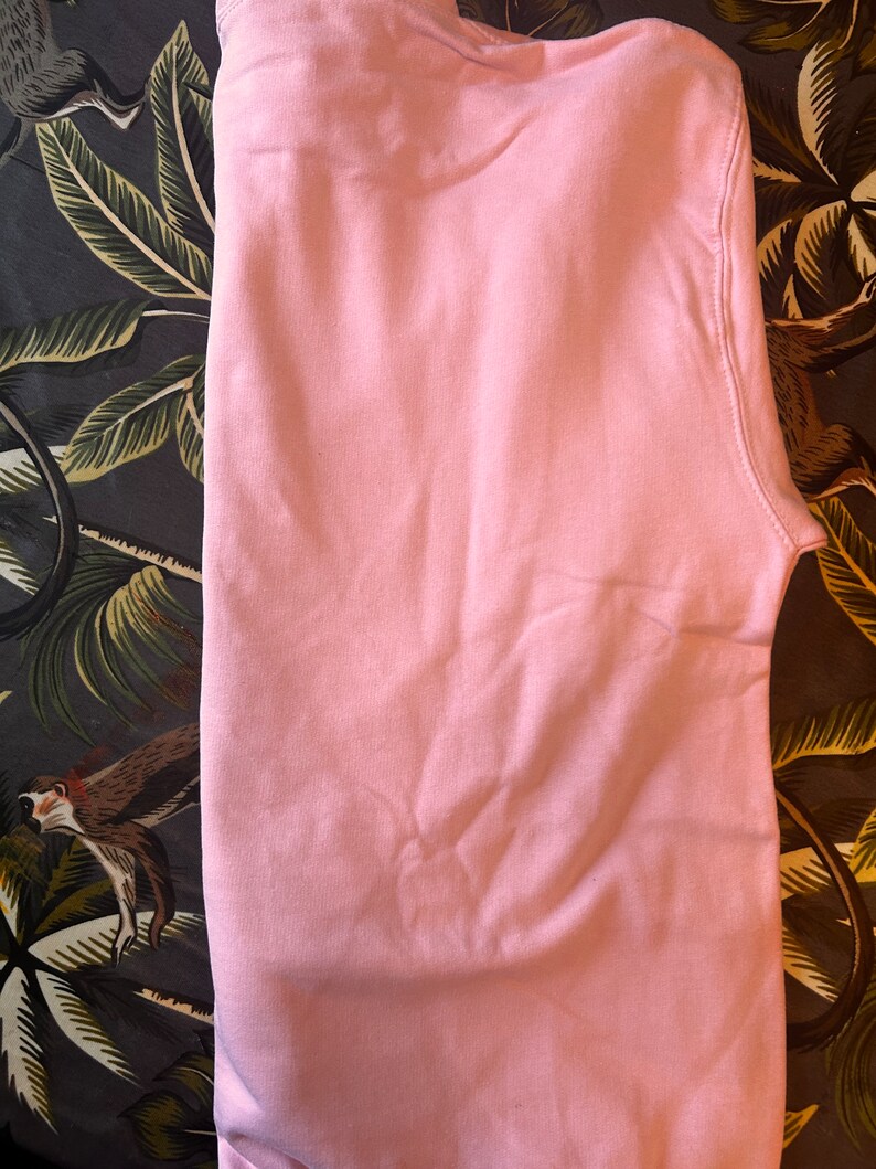 Op de afbeelding: Een effen lichtroze sweatshirt, netjes opgevouwen. De stof lijkt een zacht materiaal te zijn. De sweatshirt is effen van kleur, zonder zichtbare afbeeldingen of patronen. Het kledingstuk is waarschijnlijk voor vrijetijdsgebruik.