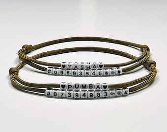 Puede incluir: Dos pulseras de cordón verde oliva con cuentas de letras blancas que deletrean "YASHA" y "PUMBA". Cada pulsera tiene una serie de cuentas numéricas y cuentas en forma de corazón. Las pulseras están sobre una superficie blanca.