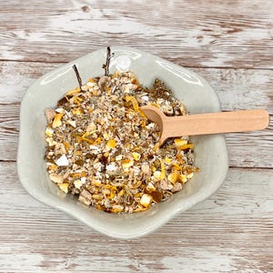 May include: A white ceramic bowl filled with a blend of dried herbs and flowers, including yellow petals, green leaves, and brown pieces. A wooden spoon rests on top of the mixture.