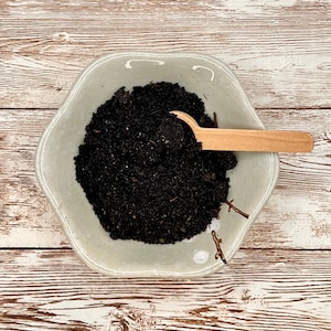 May include: A small, white ceramic bowl filled with black soil and a wooden scoop. The bowl has a decorative design on the rim.