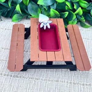 May include: A miniature picnic table with brown planks and black supports. A small, rectangular, burgundy-colored tray sits on the table. A small, white and blue decorative item is on the tray. The background features green foliage.