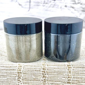May include: Two clear jars with black lids, one filled with light gold glitter and the other with dark gray glitter. The jars are cylindrical and sit on a textured, light-colored surface.