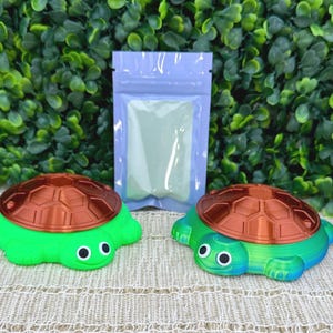 May include: Two turtle-shaped figures with copper-colored shells and googly eyes. One turtle has a solid bright green body, while the other features a blue and green gradient body. A small, light purple plastic pouch is positioned between them.