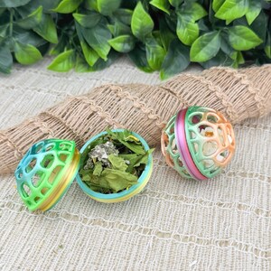 May include: Three colorful pet toy balls. One is open, revealing dried leaves inside. The balls are green, blue, yellow, pink, and orange. The toys are designed with a cage-like structure, allowing visibility of the contents.