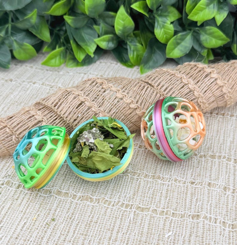 May include: Three colorful pet toy balls. One is open, revealing dried leaves inside. The balls are green, blue, yellow, pink, and orange. The toys are designed with a cage-like structure, allowing visibility of the contents.