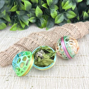 May include: Three colorful pet toys. One is a blue, green, and yellow open-design ball, another is a green, pink, and orange open-design ball, and the third is a blue and yellow bowl filled with green leaves. The toys are on a textured surface.