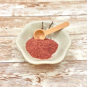 May include: A white ceramic bowl with a pink powder and a wooden spoon. The bowl has a decorative design on the side.