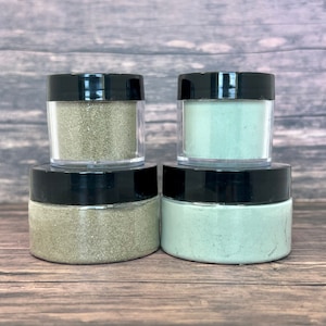 May include: Four small jars of green glitter, two are a light green and two are a darker green. The jars have black lids.